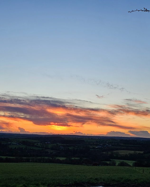 Wide view of rolling countryside at sunset, with dark green fields in the foreground and layered clouds glowing orange and gold along the distant horizon beneath a clear blue sky.