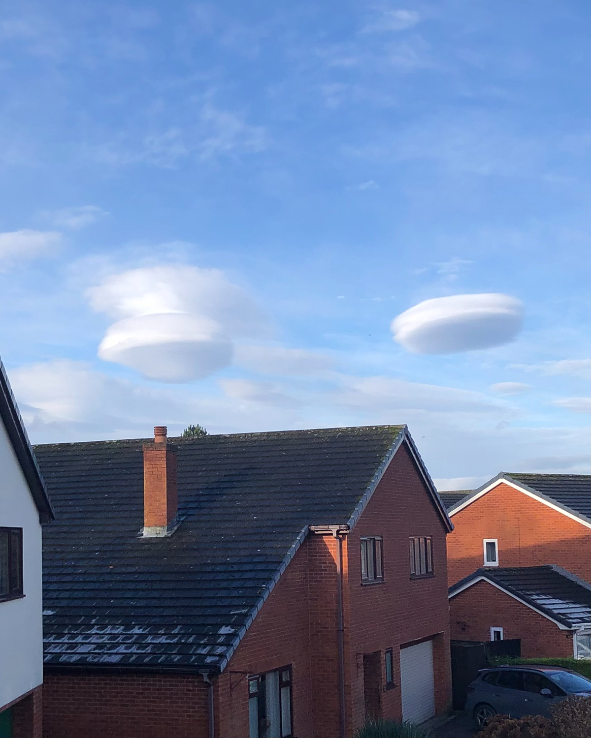 Two smooth, lens-shaped lenticular clouds floating in a blue sky above suburban brick houses with pitched roofs, chimneys, and parked cars visible below.