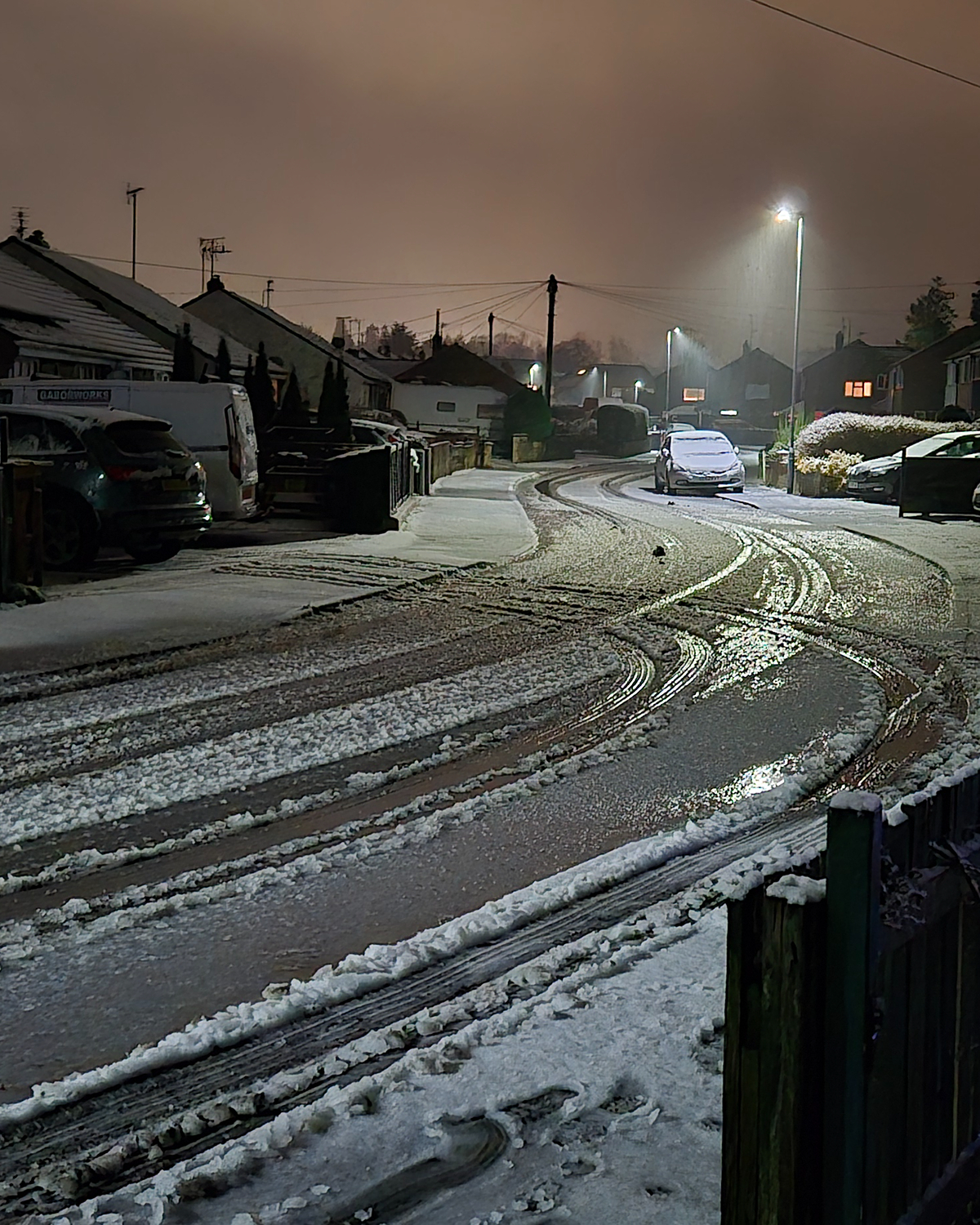 Snowy residential street at night with icy, slushy road surfaces and visible tyre tracks, parked cars along the curb, and streetlights reflecting off the wet ice under an overcast sky.