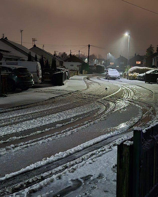 Snowy residential street at night with icy, slushy road surfaces and visible tyre tracks, parked cars along the curb, and streetlights reflecting off the wet ice under an overcast sky.