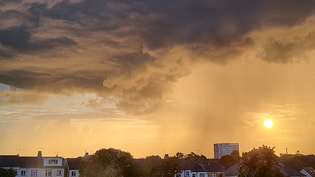 Dark storm clouds hang over a town at sunset, with streaks of rain falling from the clouds while the sun shines brightly through the golden sky.
