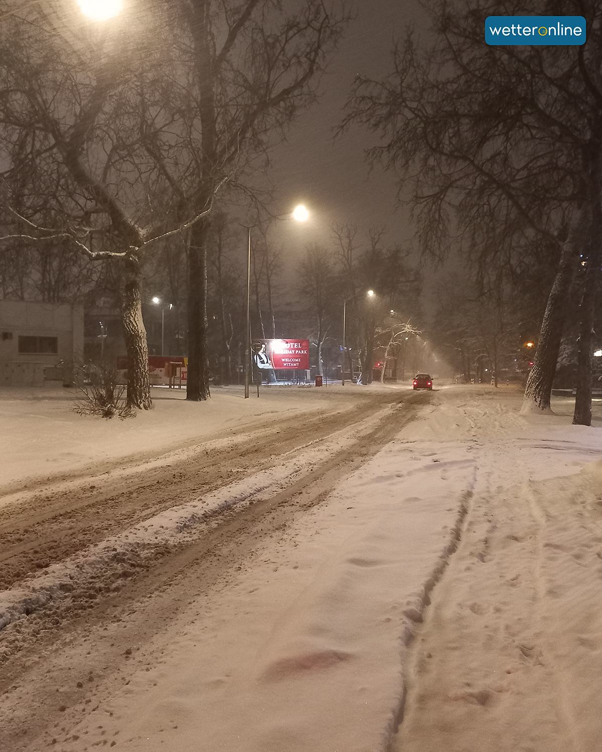 Breite Straße mit Schnee, Bäumen und Straßenlaternen in winterlicher Nachtstimmung.