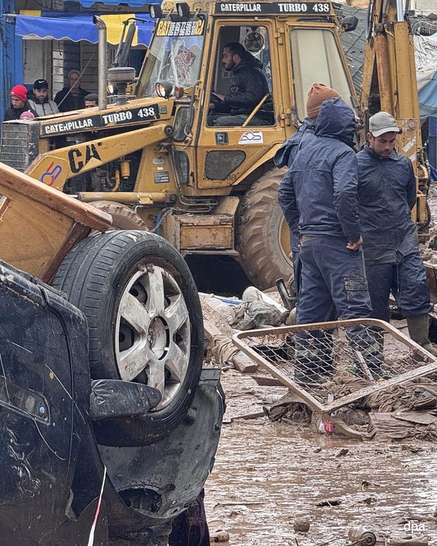 Les secours et une pelleteuse déblayent une zone inondée et détruite où se trouvent des voitures renversées. La boue et les débris recouvrent la route.