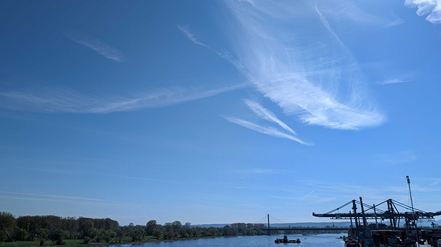 Flusslandschaft mit Hafenanlagen unter blauem Himmel mit dünnen Schleierwolken.