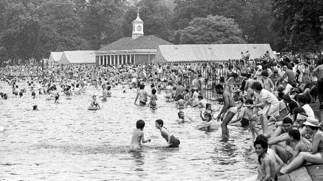 Black-and-white photograph from the 1976 UK heatwave showing a crowded outdoor swimming area packed with people bathing, wading, and sitting along the water's edge during extreme summer heat.