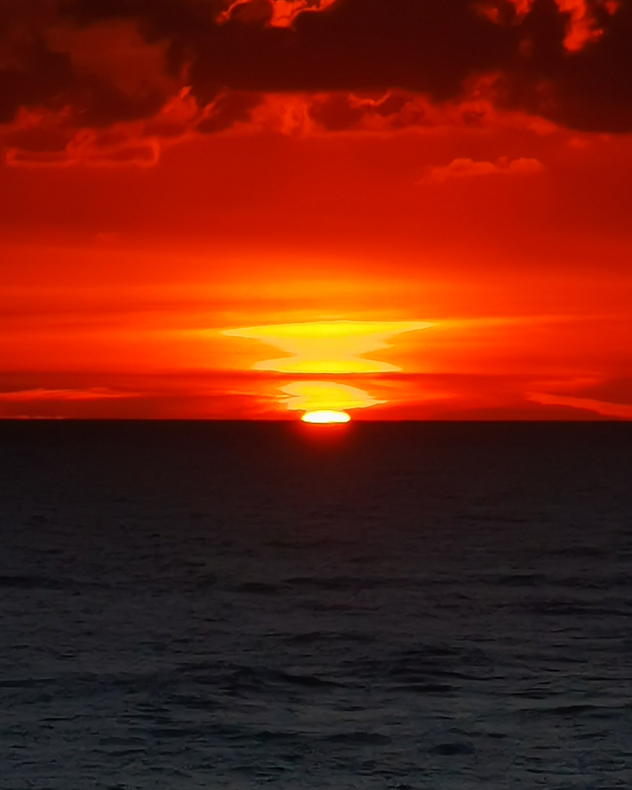 Brilliant red-orange sunset over a dark sea, with the sun touching the horizon beneath glowing layered clouds and a dark cloud bank.
