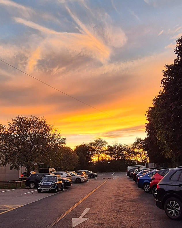 Golden-orange sunset illuminates wispy, swirling clouds above a car park lined with vehicles and trees, with a blue-gray evening sky.