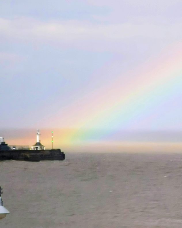 Faint pastel rainbow arching over a gray sea beside a small lighthouse and harbor wall, with soft pink, green, and blue colors visible against a misty sky.