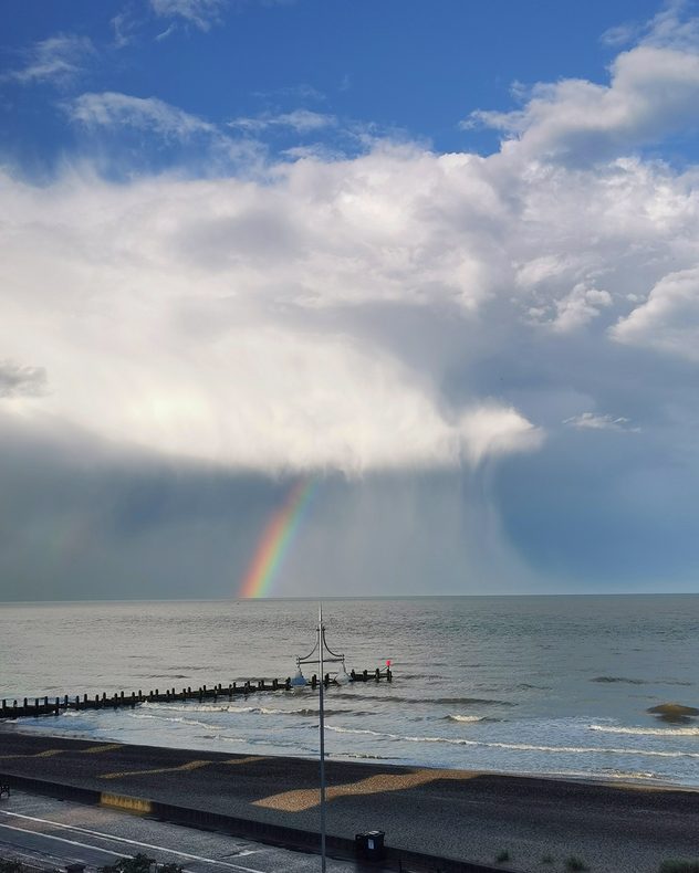 Coastal scene with a vivid rainbow descending from a towering rain cloud over the sea, visible rain shafts reaching the water, and gentle waves breaking along the shoreline.