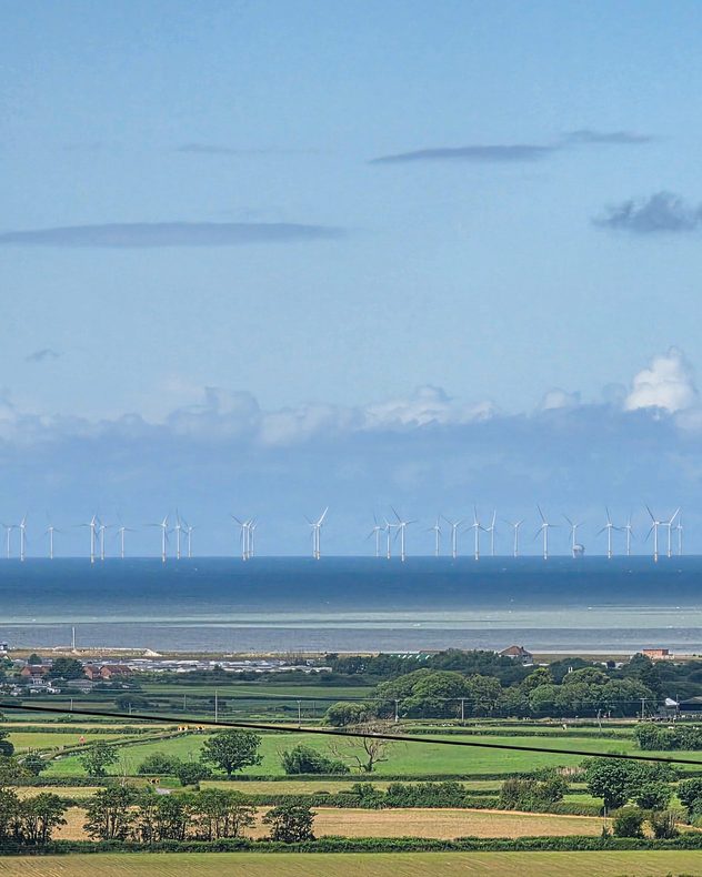 Distant offshore wind turbines stretching across the horizon above a calm blue sea, viewed from rolling green farmland and coastal countryside under layered clouds.