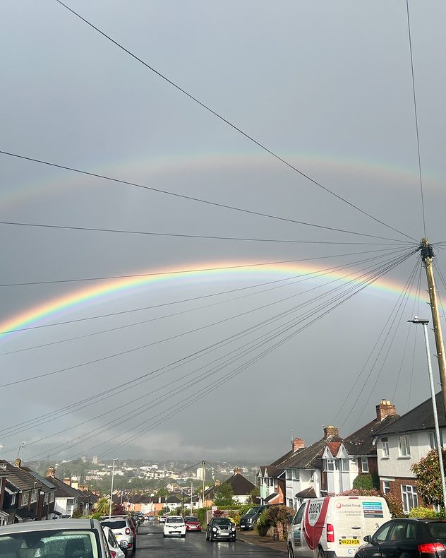 Bright double rainbow arching across a gray stormy sky above a residential street, with wet roads, parked cars, rooftops, and utility wires converging toward a central pole.