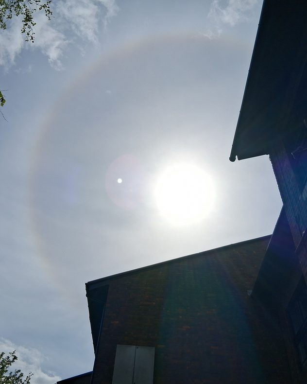 Bright sun shining through a thin veil of high cloud above brick rooftops, with a faint circular halo around the sun and lens flare visible in the sky.