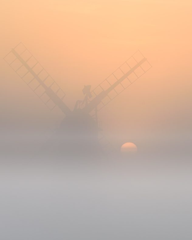 Traditional windmill silhouette partly hidden by dense fog at sunrise, with a pale orange sun low on the horizon beneath a soft peach-colored sky.