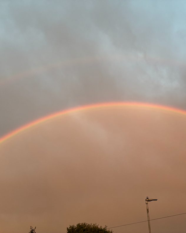 Bright rainbow arching across a gray stormy sky with soft orange light beneath the clouds and a streetlight silhouette in the foreground.