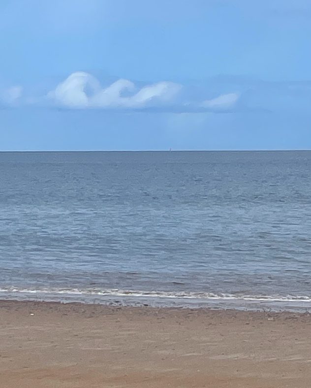 Calm beach and flat sea beneath unusual rolling wave-shaped clouds aligned along the horizon under a pale blue sky.