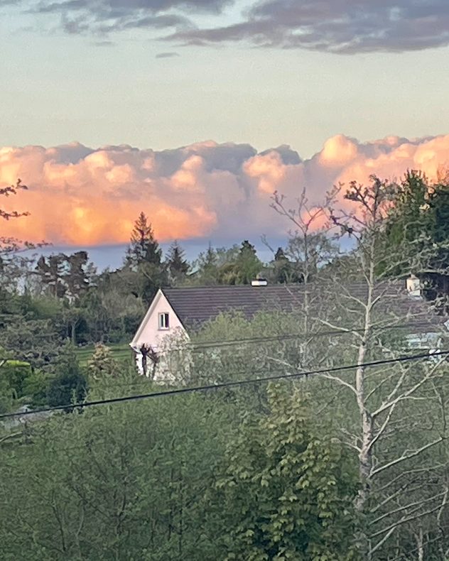 Large pink-orange sunset clouds above a rural neighborhood with trees and rooftops, illuminated by warm evening light beneath darker gray clouds.