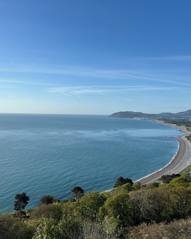 Wide coastal bay with calm blue water, a curving shoreline, and distant green hills beneath a clear blue sky viewed from an elevated hillside.