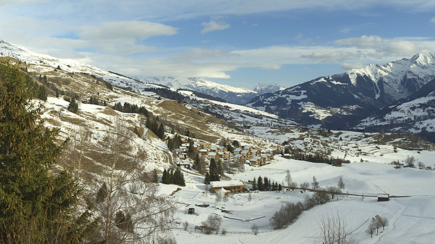 Teils verschneite Alpenlandschaft mit einem kleinen Dorf, sanften Hängen und umliegenden Bergen unter wechselnder Bewölkung.