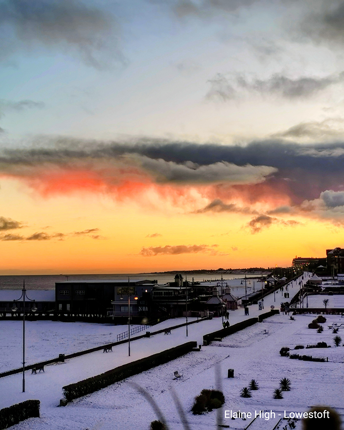 Promenade côtière enneigée en Angleterre avec des chemins, des bancs et des bâtiments. En arrière-plan, la mer et, au-dessus, un ciel coloré au coucher du soleil.