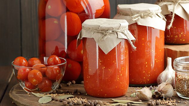 Eingekochte Tomaten in Gläsern mit Stoffabdeckung, daneben frische Kirschtomaten, Knoblauch und Gewürze auf einem Holztisch. (c) teatian/Shutterstock.com