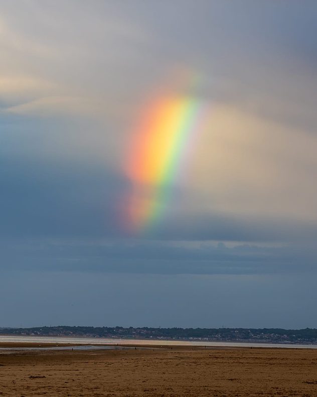 Bright partial rainbow descending from grey rain clouds above a wide tidal shoreline, with exposed sand flats, calm water, and a distant coastal town along the horizon.