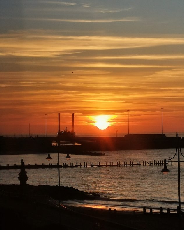 Fiery orange sunset over a coastal harbour with calm water, silhouetted piers, lamp posts, and industrial structures along the horizon beneath layered evening clouds.