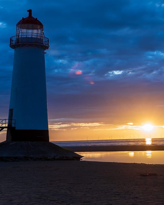 White lighthouse silhouetted against a golden sunset over the sea, with dark blue clouds overhead, reflections on calm water, and distant offshore wind turbines along the horizon.