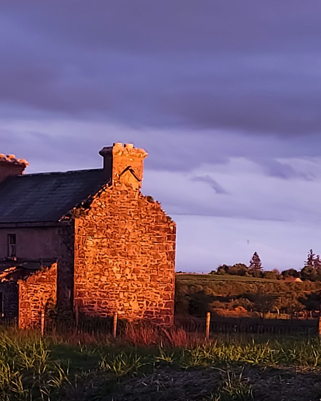 Stone cottage illuminated by warm golden sunset light beside grassy fields, with dark purple-grey storm clouds overhead and a calm coastal horizon in the horizon.