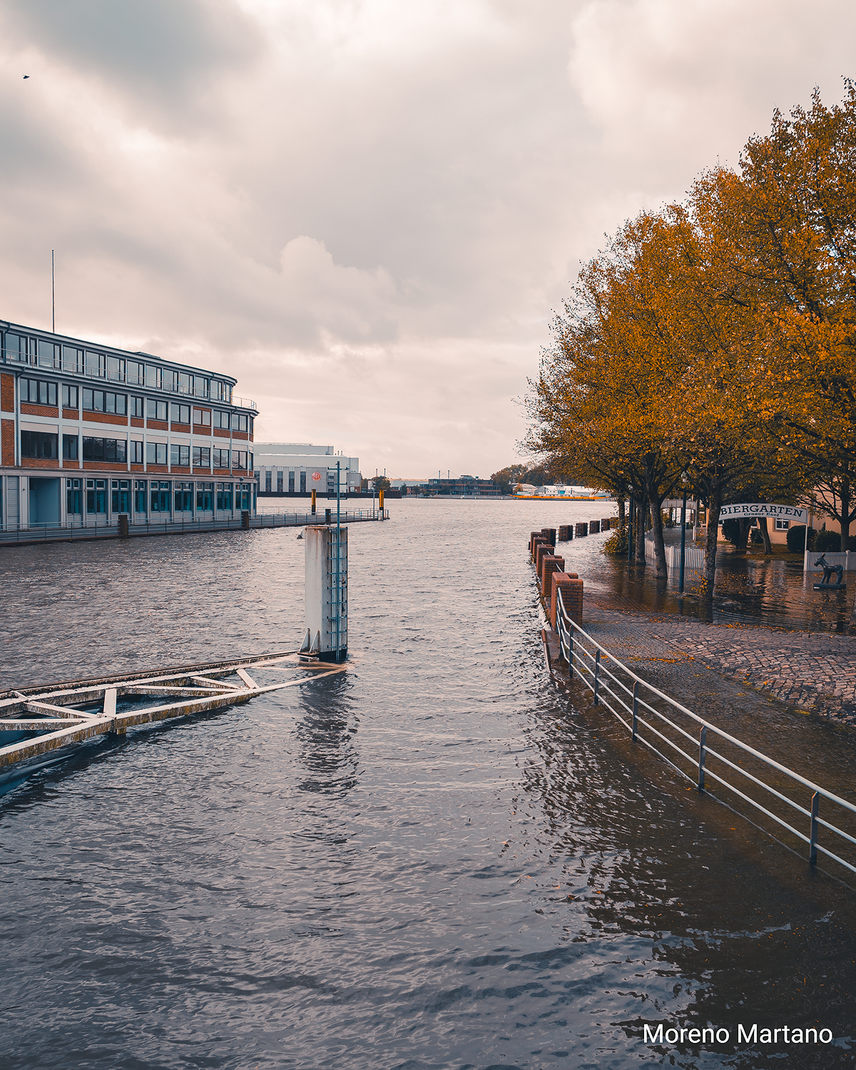 Überflutetes Weserufer in Bremen, Wasser reicht bis an Gehwege und Bäume, im Hintergrund Gebäude unter grauem Himmel.