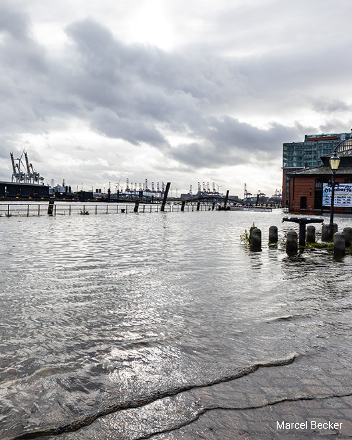 Überfluteter Hamburger Fischmarkt, Wasser bedeckt die gepflasterte Fläche, im Hintergrund Hafenkräne und Speicherstadt-Gebäude.