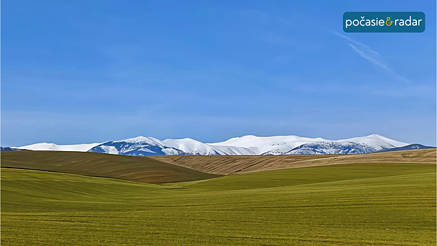 Zelená krajina s poľami a zasneženými horami na horizonte pod jasnou modrou oblohou