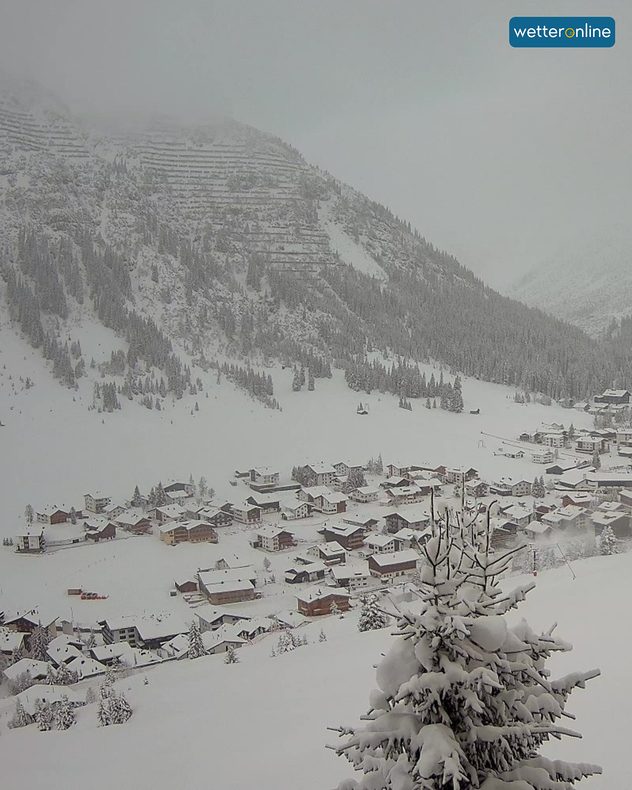 Verschneites Alpendorf mit wolkenverhangenen Bergen und schneebedeckter Tanne im Vordergrund.