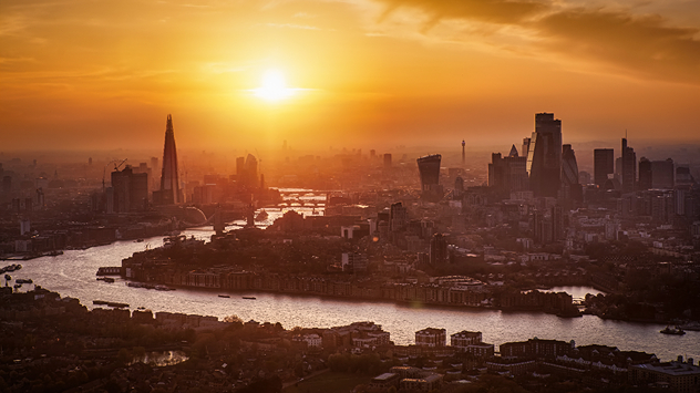 Aerial view of the London skyline at sunset with the River Thames winding through the city, the Shard and City skyscrapers silhouetted in hazy golden light, highlighting hot conditions.