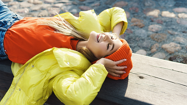 Junge Frau im farbenfrohen Outfit genießt die Sonne auf einer Bank liegend.