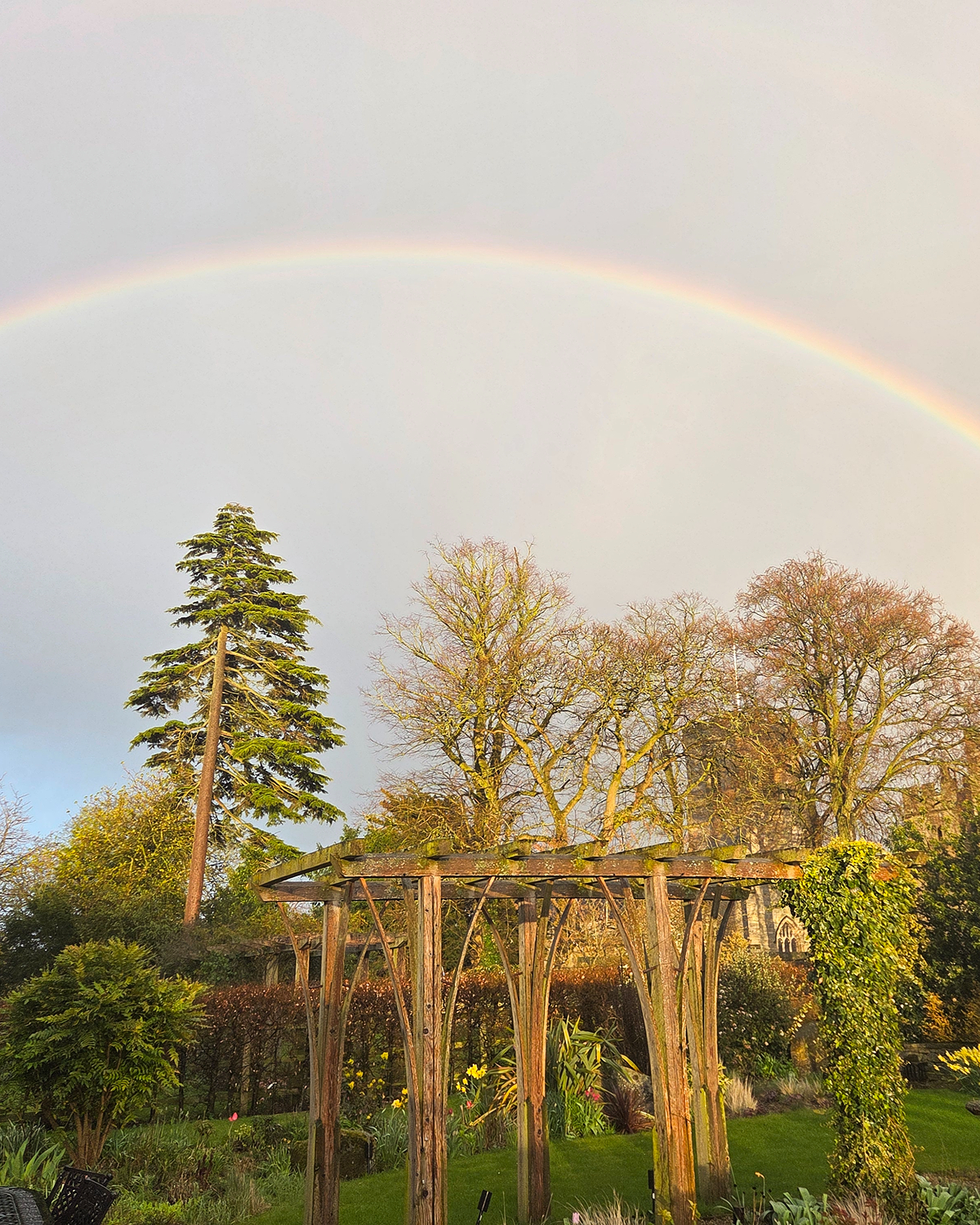Faint rainbow arching across a pale sky above a garden with a wooden pergola, green lawn, and trees lit by warm sunlight.