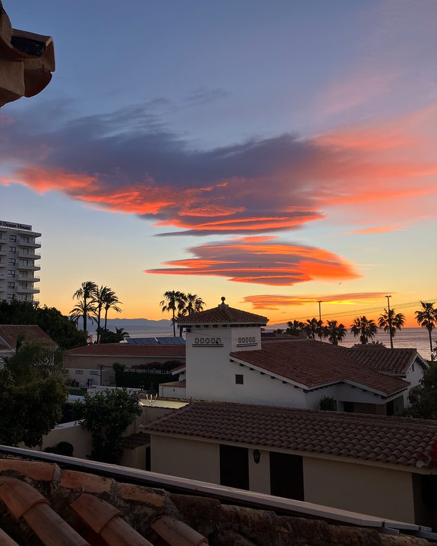Coastal sunset over seaside buildings with palm trees, featuring layered lenticular clouds glowing orange and pink above the horizon and calm sea in the background.