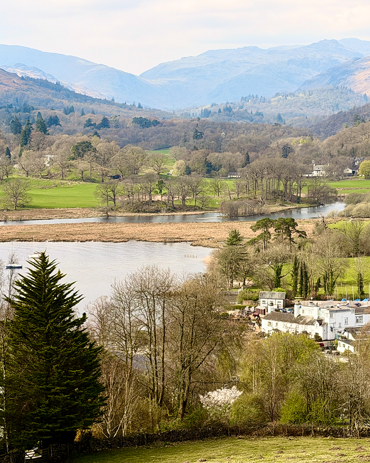 Scenic valley with a winding river or lake, surrounded by green fields, trees, and layered hills fading into the distance under a soft hazy sky.