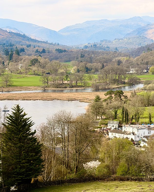 Scenic valley with a winding river or lake, surrounded by green fields, trees, and layered hills fading into the distance under a soft hazy sky.