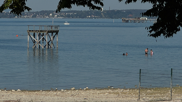 Badegäste stehen im flachen Wasser vor einer freigelegten, steinigen Uferzone und einem Steg.
