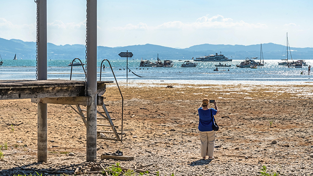 Eine Frau fotografiert das weit zurückgewichene Seeufer; im Hintergrund liegen zahlreiche Boote auf dem Wasser.