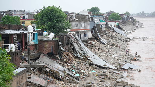 © dpa Häuser am Ufer sind durch Überschwemmungen und Erdrutsche schwer beschädigt, Trümmer liegen verstreut, Menschen gehen am Fluss entlang.