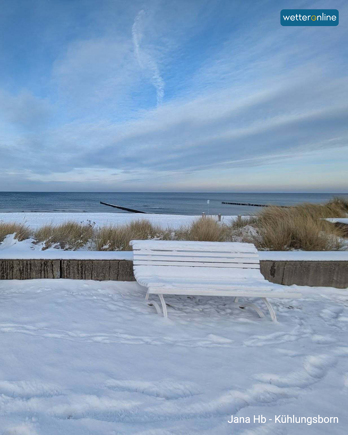 Verschneite Parkbank am Strand von Kühlungsborn, dahinter ruhige Ostsee und blauer Winterhimmel.