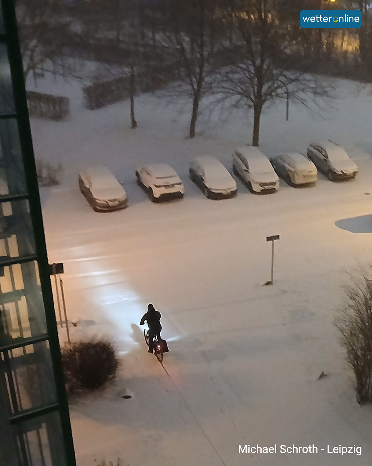Person mit Fahrrad bei Schneefall auf verschneitem Parkplatz.