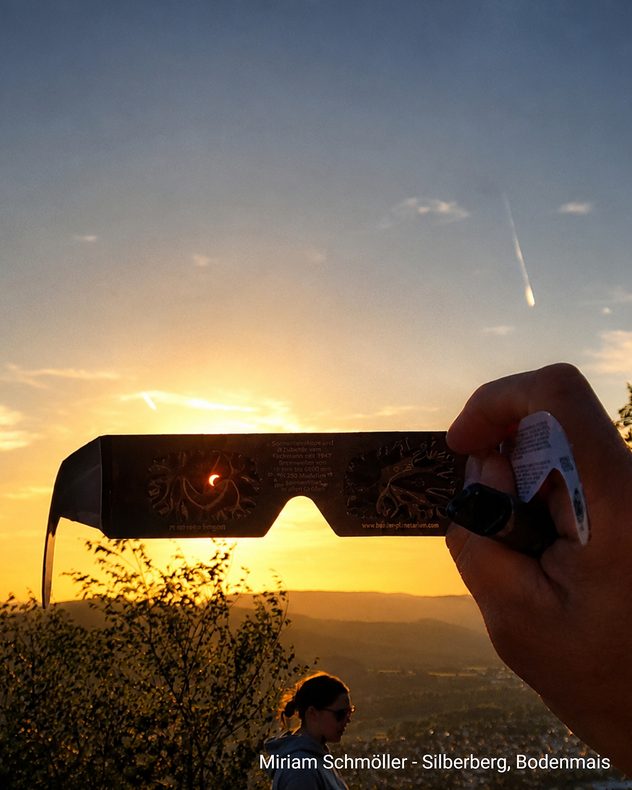 Eine Hand hält eine Finsternisbrille vor den Abendhimmel. Durch sie ist die teilweise verdeckte Sonne zu sehen.