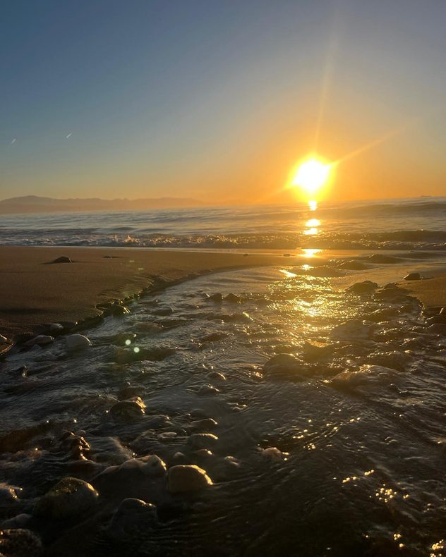 Golden sunrise over the sea with a small stream flowing across a sandy beach, reflecting bright sunlight and illuminating pebbles and gentle waves.