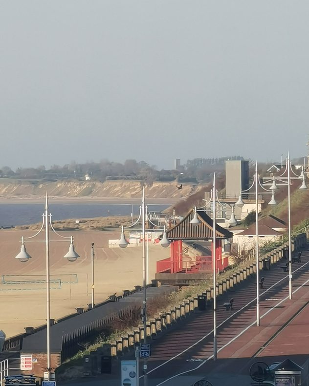 Coastal promenade with decorative lampposts and a red shelter beside a sandy beach, calm sea and distant cliffs under a pale hazy sky.