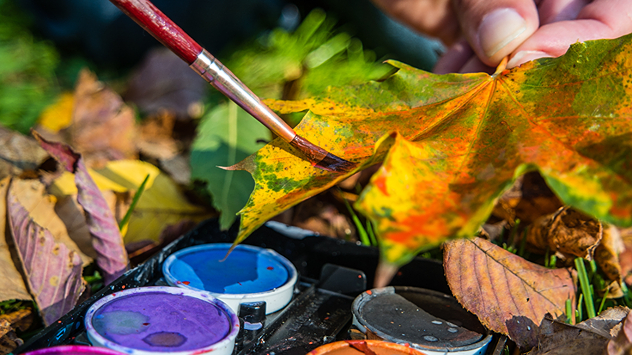 Ein herbstlich gefärbtes Ahornblatt wird mit Wasserfarben bemalt – im Vordergrund liegt ein Farbkasten zwischen buntem Laub.