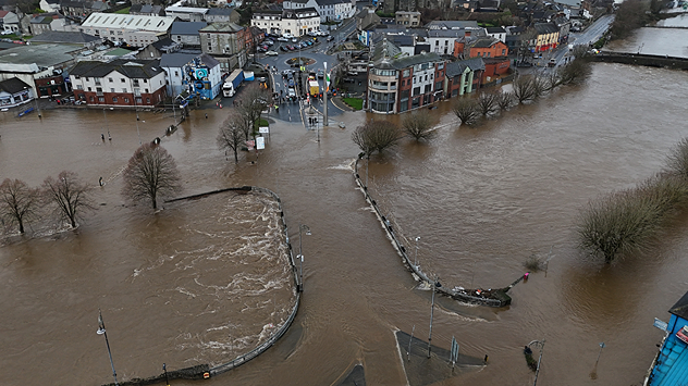 Aerial view of a town with rivers overflowed, brown floodwater covering roads, parks, and footpaths, surrounding buildings and trees.
