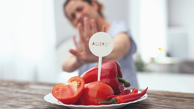 Teller mit rohem Gemüse (Tomaten, Paprika, Chili) und „Allergy“-Hinweis, im Hintergrund eine Frau mit abwehrender Geste.
