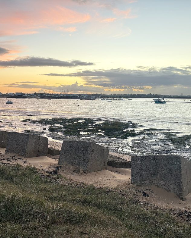 Coastal estuary at low tide with exposed mudflats, anchored boats, concrete blocks in the foreground, and a pastel orange-pink sunset behind distant buildings.